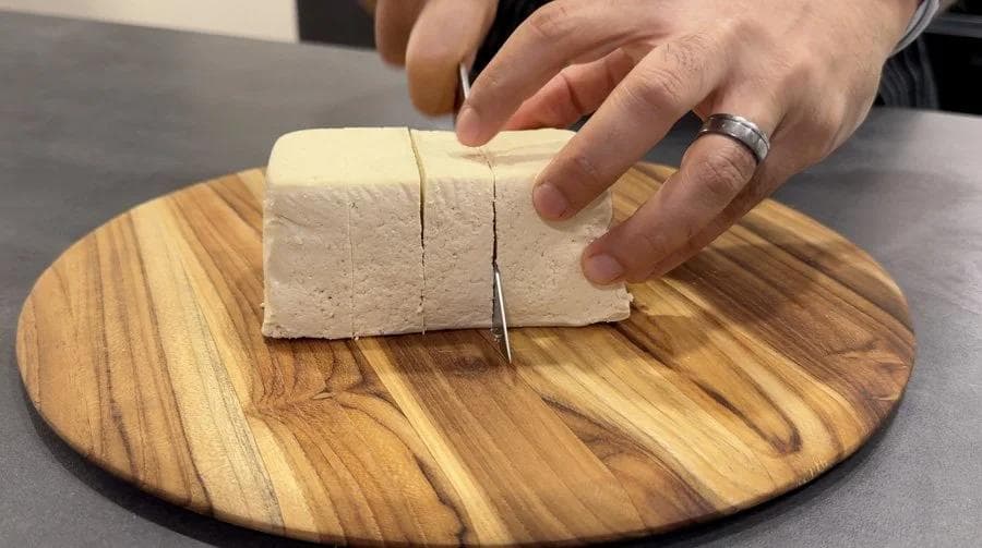 Hands cutting pressed tofu into cubes on a wooden cutting board with a knife.