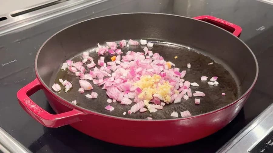 Pink and yellow diced onions and garlic cooking in olive oil in a red cast-iron pan on stovetop.