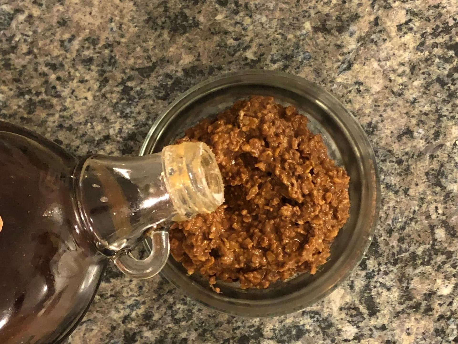 Maple syrup being drizzled from a glass bottle over a bowl of chocolate-coated oatmeal on a granite countertop.