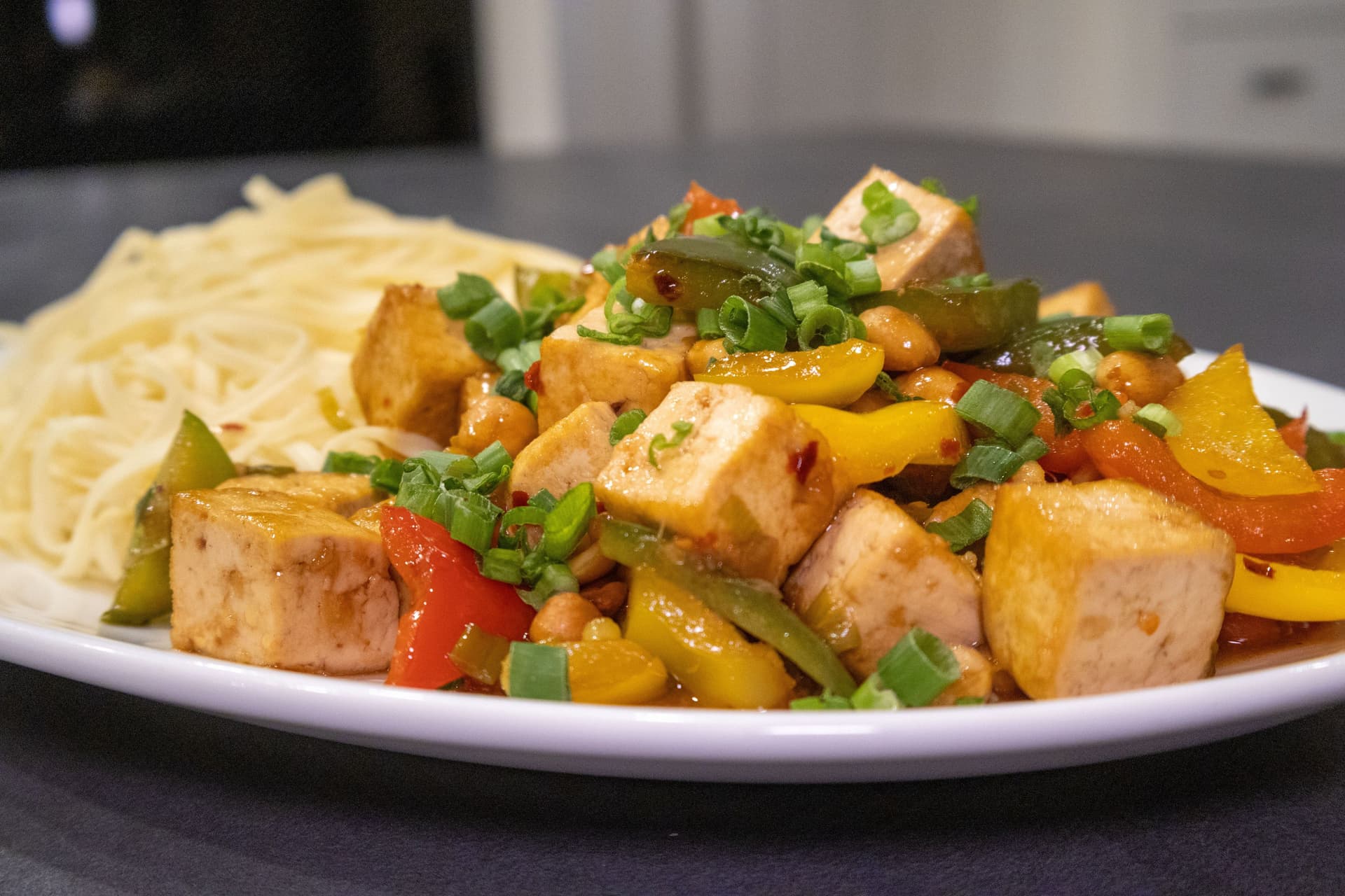 Tofu noodle stir fry with cubed tofu, bell peppers, and green onions served over thin noodles on a white plate.