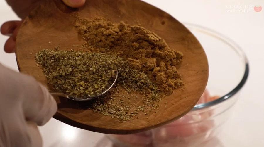 Spoon holding a mixture of dried oregano and cumin over a wooden plate with ground spices for Cuban mojo chicken marinade.