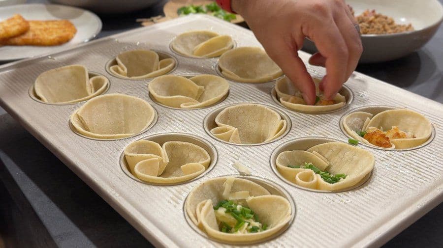 Tortilla egg cups arranged in a muffin tin, some filled with vegetables and cheese, others empty, being prepared for baking.