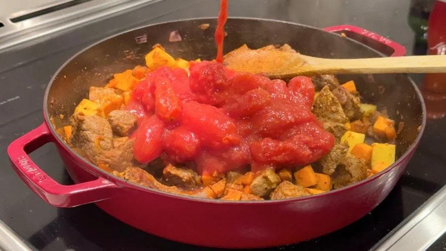 Red tomatoes being poured into a pink Dutch oven filled with beef stew ingredients and diced vegetables.