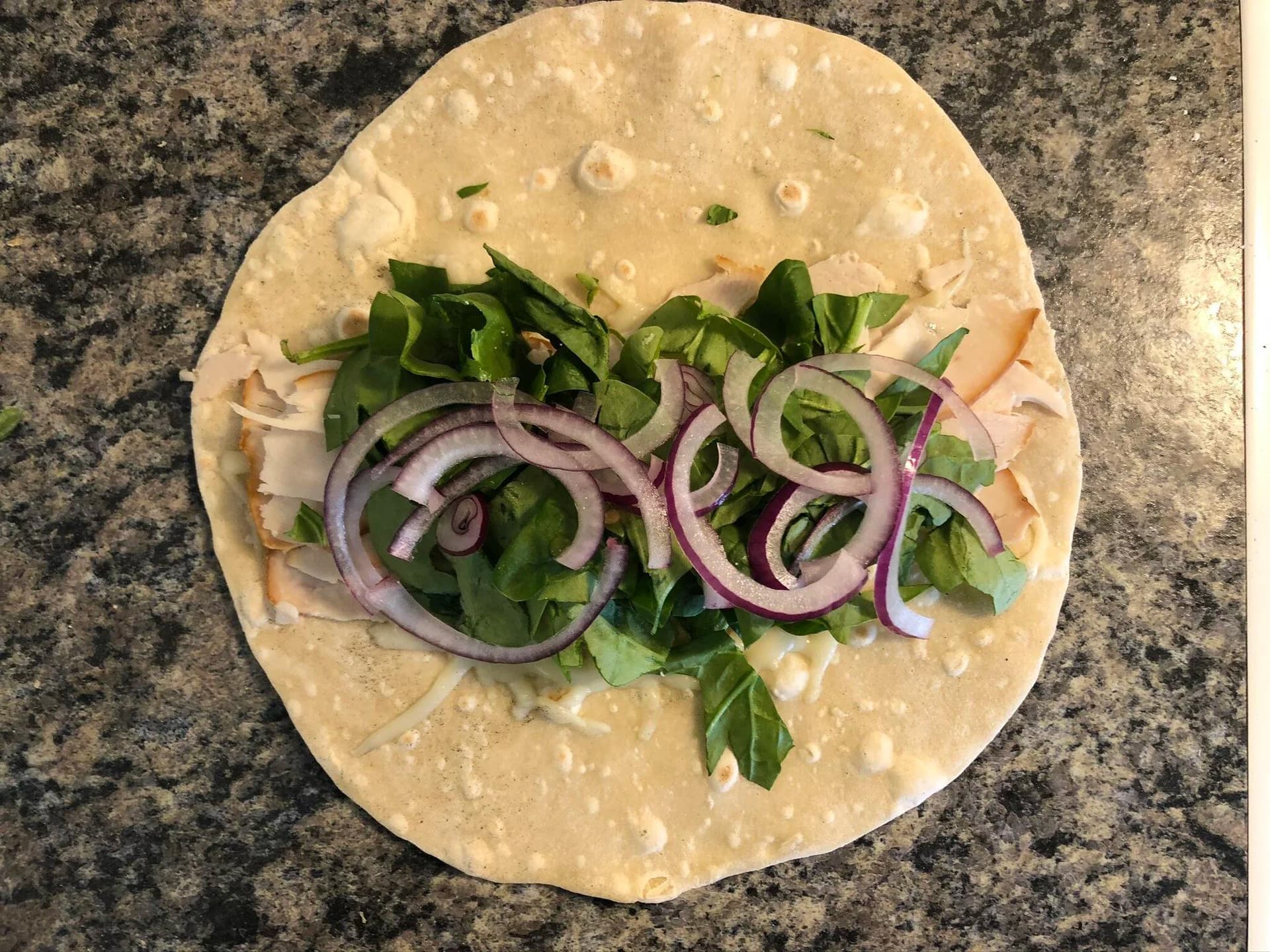 Warm tortilla topped with sliced deli turkey, fresh spinach, and purple onion rings on a granite countertop.