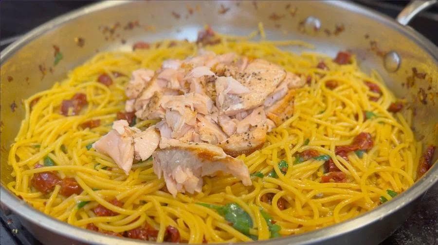 Lemon spaghetti with flaked salmon, red peppers, and green garnish in a pan, seasoned with black pepper.