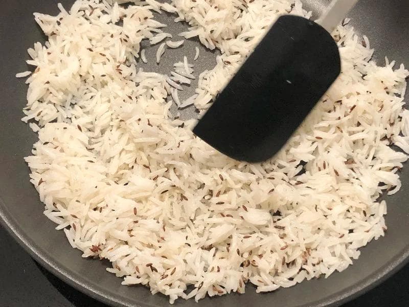 Cooked rice being stirred with a black spatula in a gray pan after oil and cumin seeds have been added.