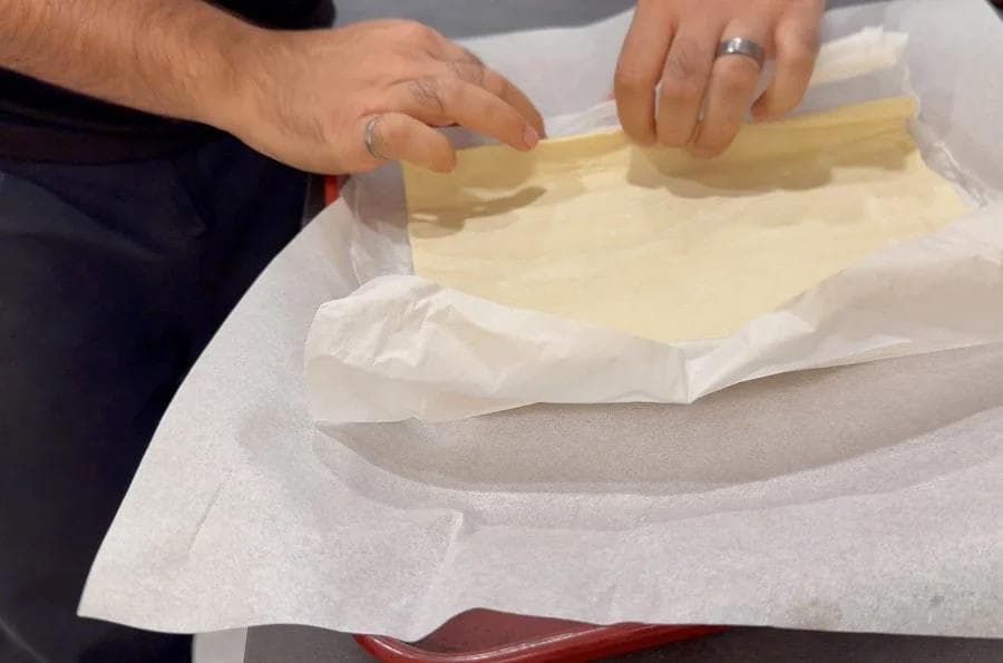 Hands folding the edges of puff pastry on parchment paper over a baking sheet.