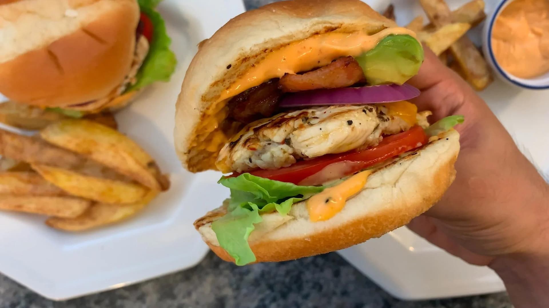 Hand holding a grilled chicken burger with cheese, tomato, lettuce, and red onion on a toasted bun, with fries on the side.