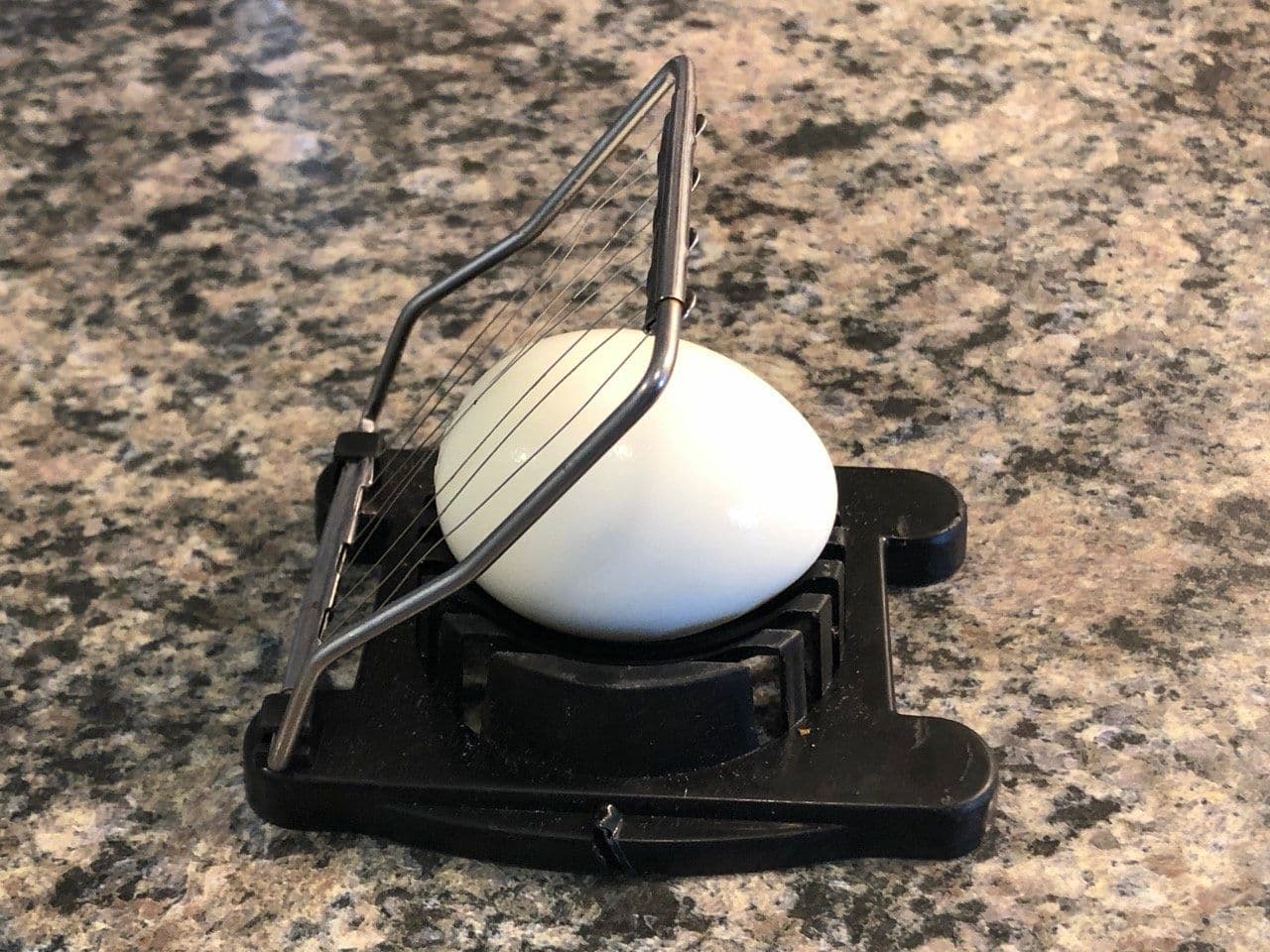 Hard-boiled egg being sliced in a black egg slicer with metal wires on a granite countertop.