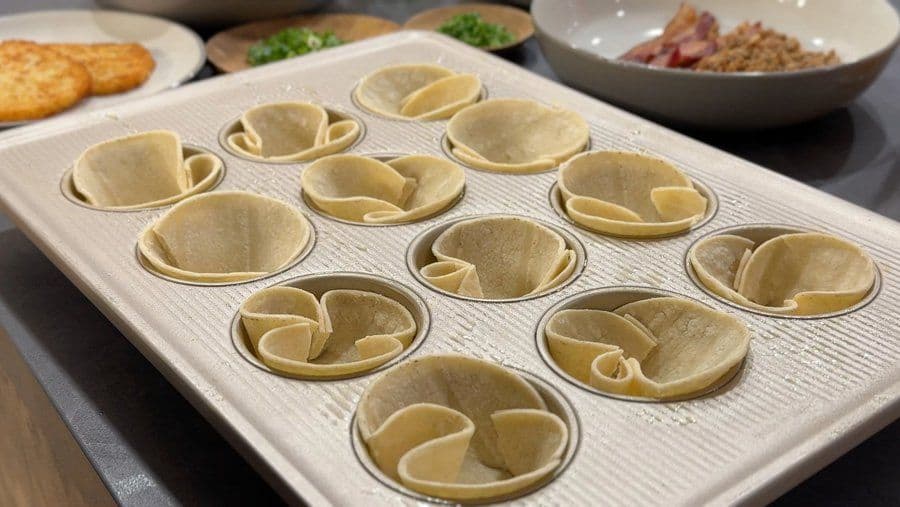 Flour tortillas pressed into muffin cups, folded to create egg cup shapes in a white muffin tin.
