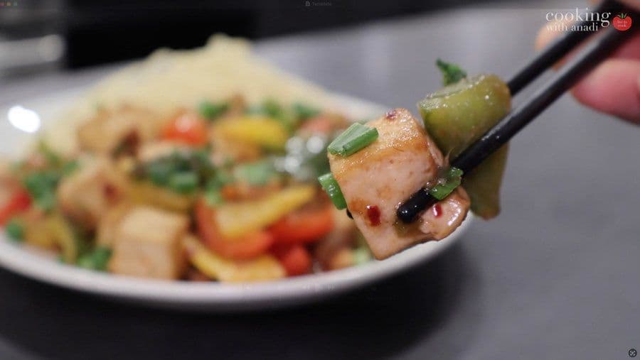 Tofu noodle stir fry held on chopsticks with green onion garnish and sauce coating, blurred plated dish in background.