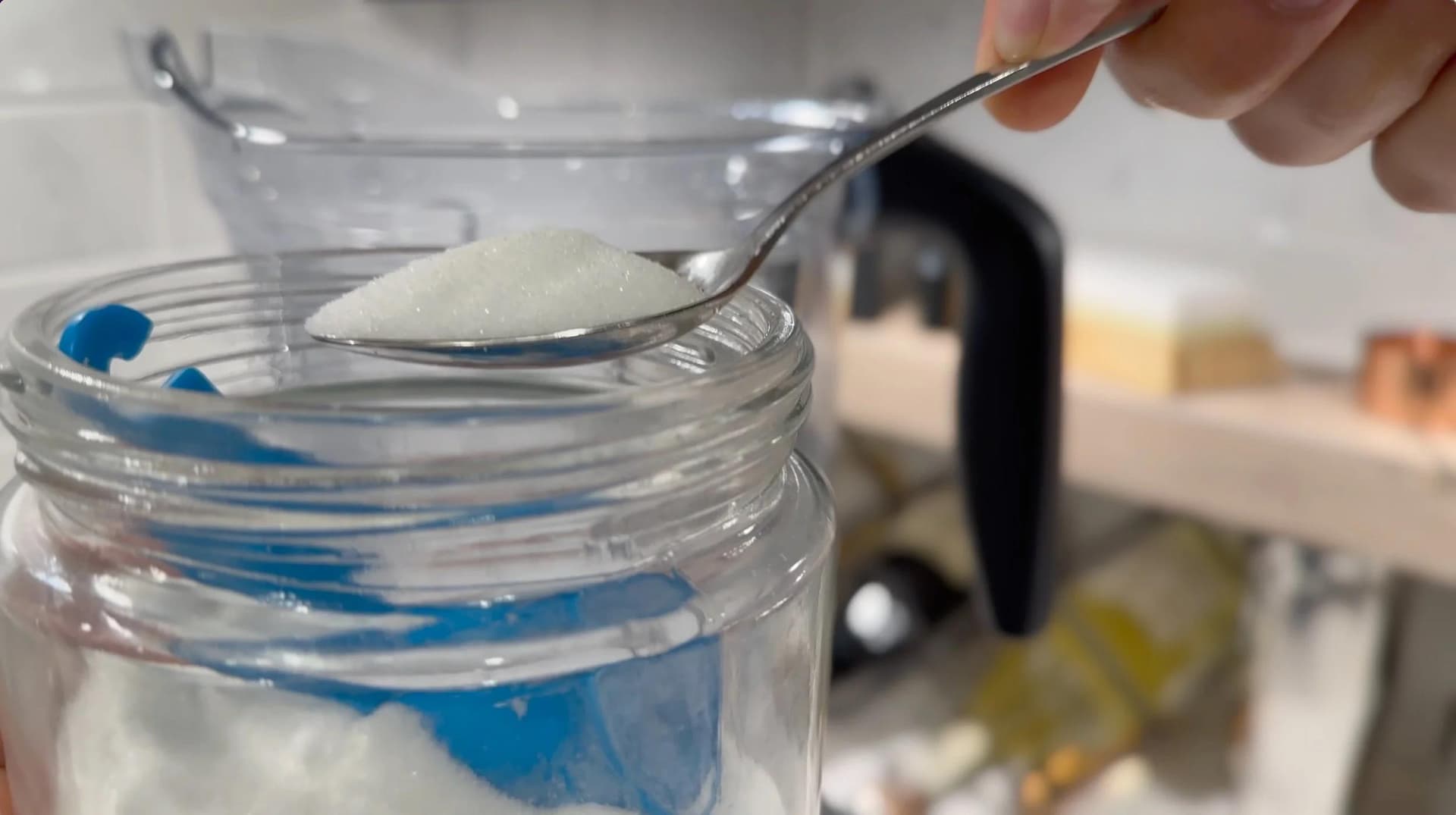 Spoon holding measured white sugar over a glass jar of blue-striped liquid in a kitchen setting.