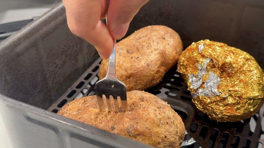 Hand using a fork to test doneness of air-fried potatoes wrapped in foil in an air fryer basket.