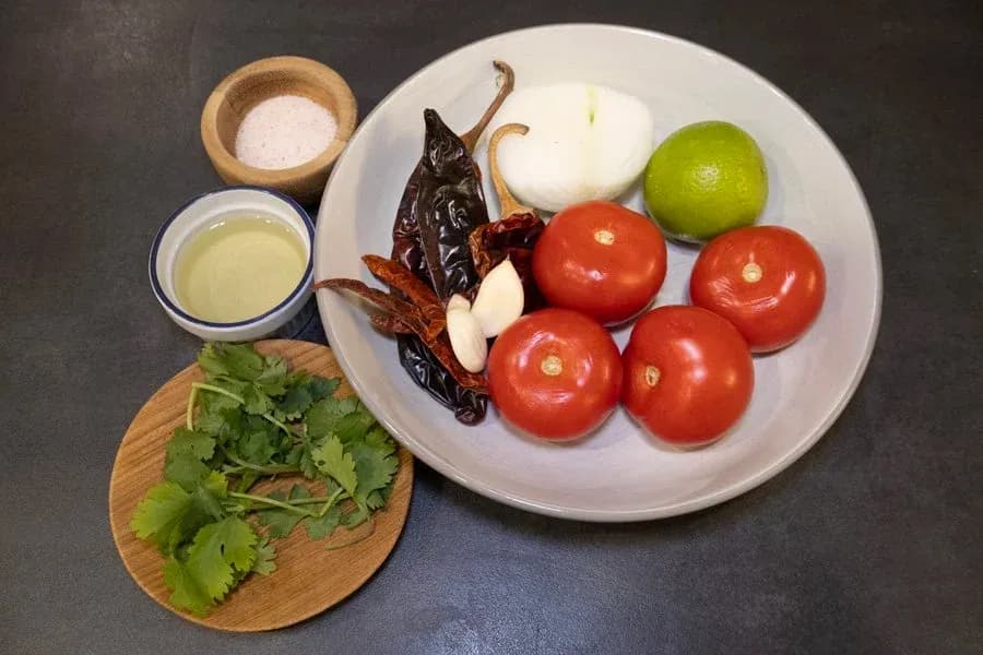 Salsa Roja ingredients arranged on a dark surface: tomatoes, dried chilies, onion, garlic, lime, cilantro, salt, and oil.