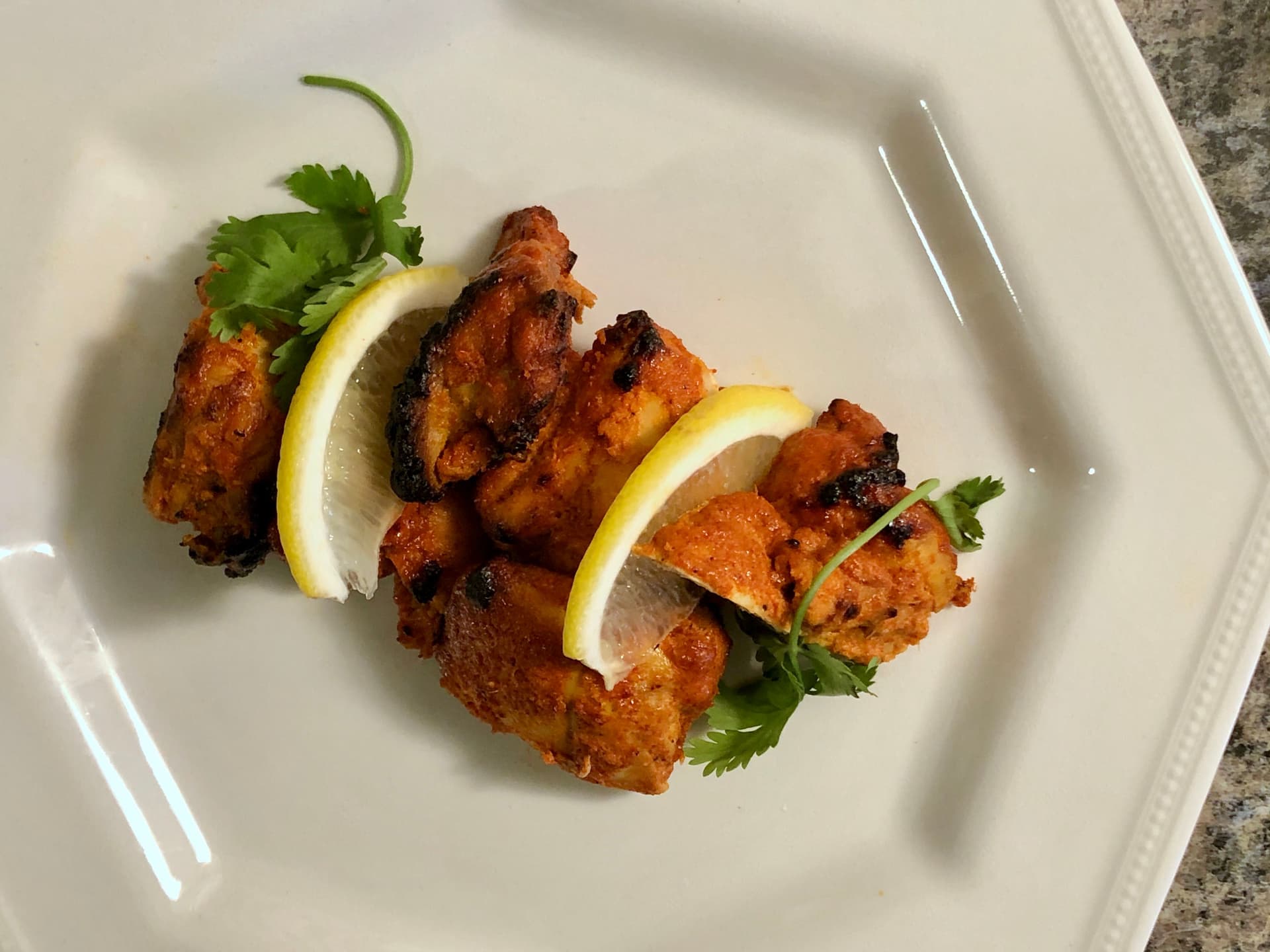 Baked chicken tikka pieces with charred edges, garnished with lemon slices and fresh cilantro on a white plate.