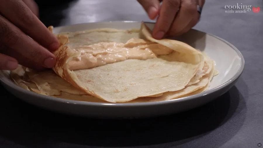 Spreading pumpkin cream cheese filling down the center of a cooked crepe on a white plate.