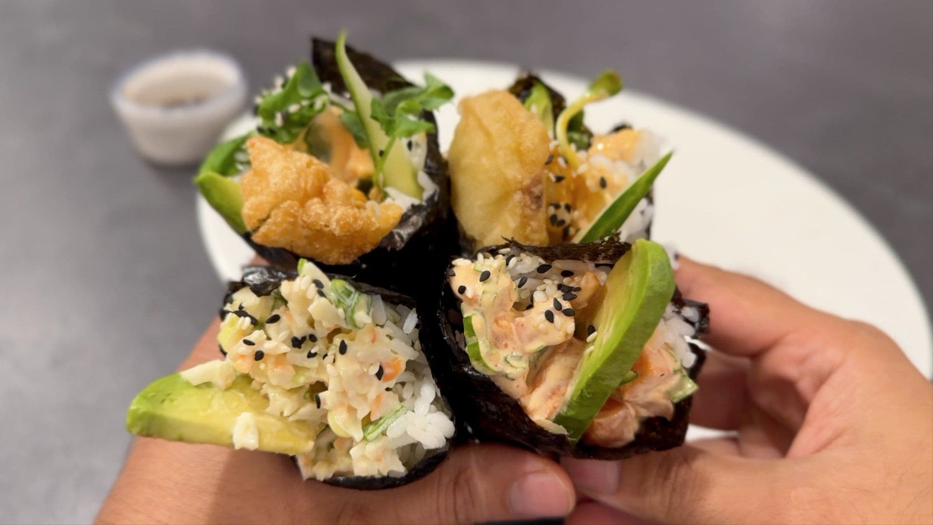 Hand holding four sushi hand rolls (temaki) with nori, rice, avocado, cucumber, and sesame seeds.