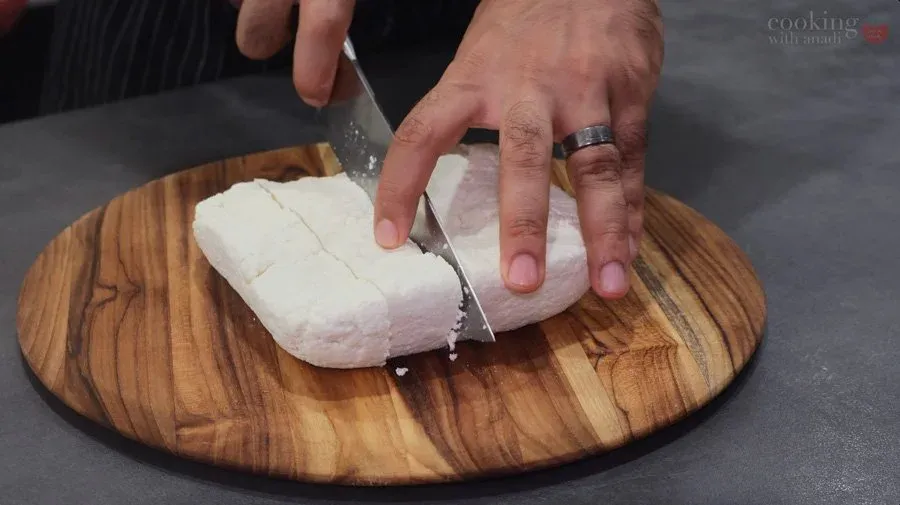 Hands slicing white paneer cheese into cubes on a wooden round cutting board with a knife.