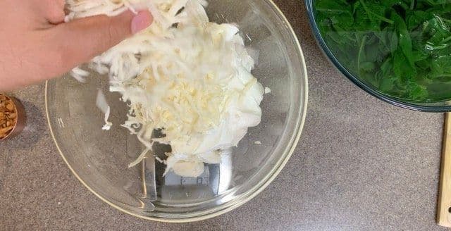 Hand holding shredded mozzarella cheese over a glass bowl with creamy dip ingredients mixed together.