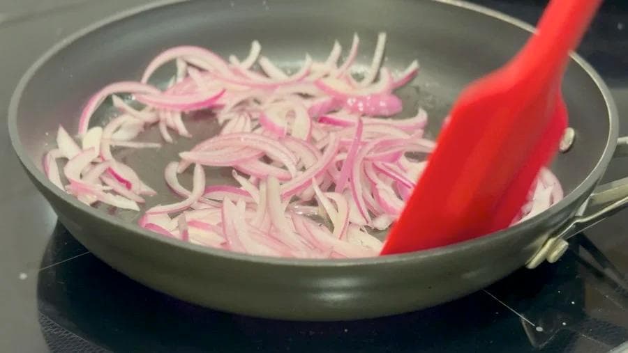 Sliced pink onions being sautéed in a pan with a red spatula on a stovetop.