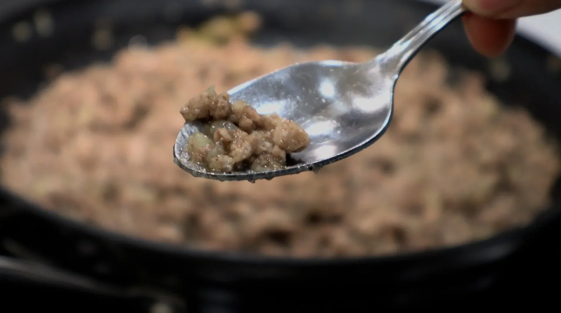 Spoon holding a portion of cooked ground meat filling with potatoes and spices over a skillet of tourtiere mixture.
