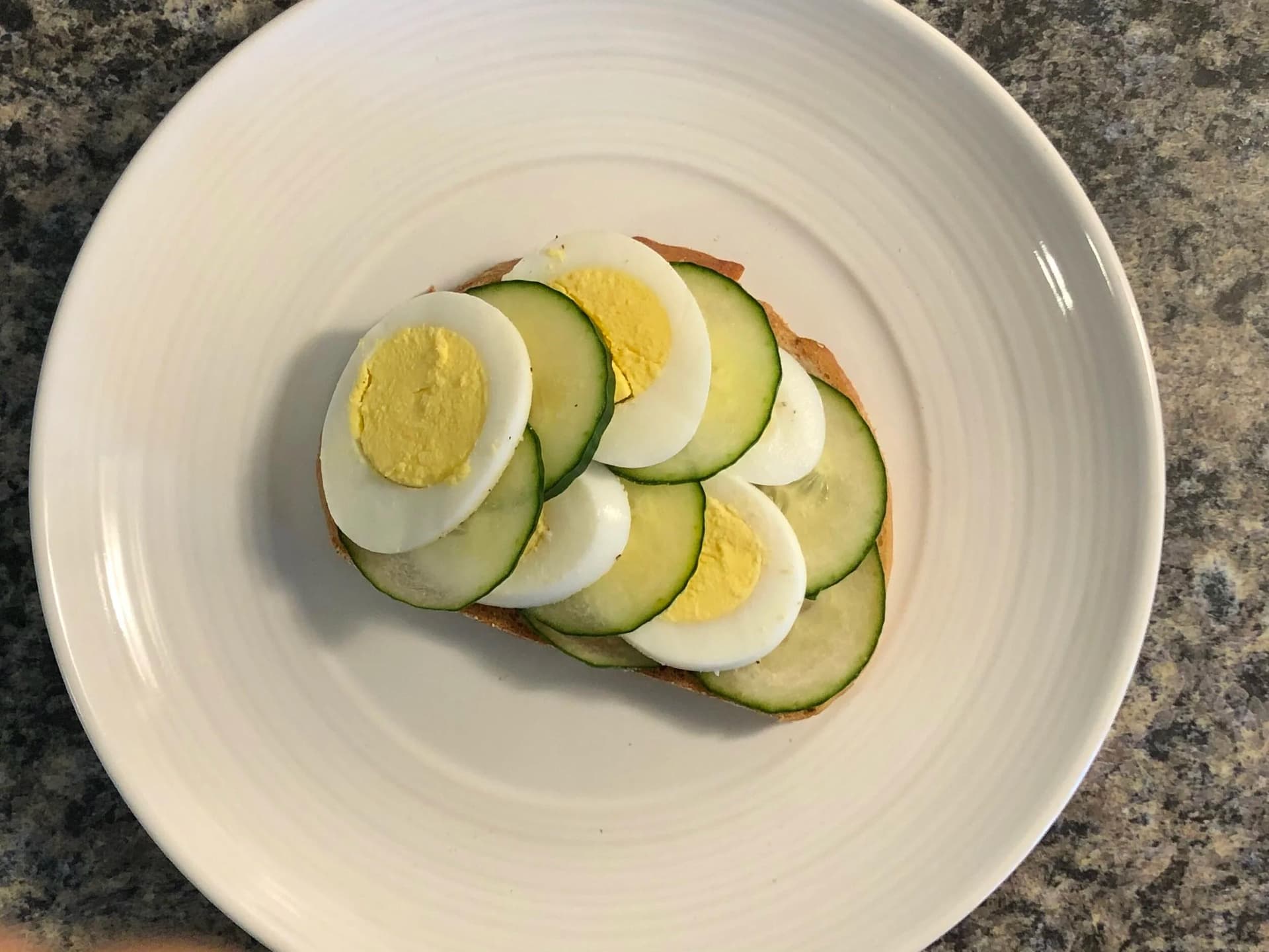 Cucumber and hard-boiled egg open-faced sandwich on toasted bread arranged on a white plate.