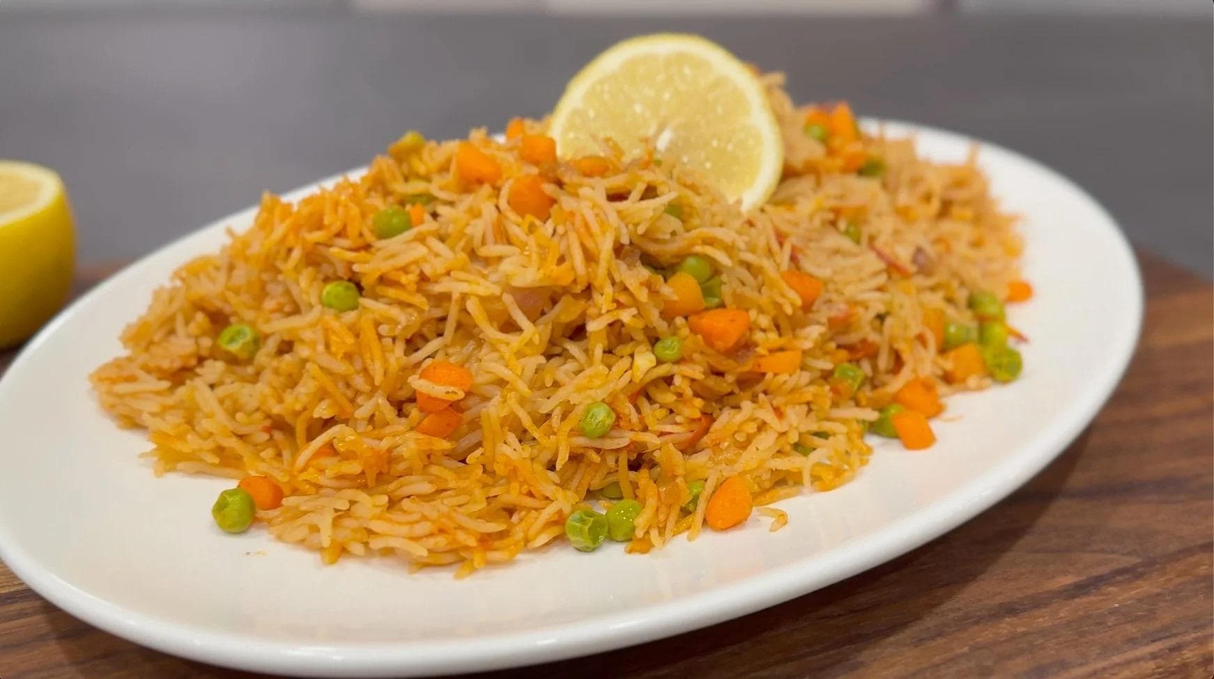 Fluffy Mexican tomato rice garnished with lemon slice, topped with peas and diced carrots on a white plate.