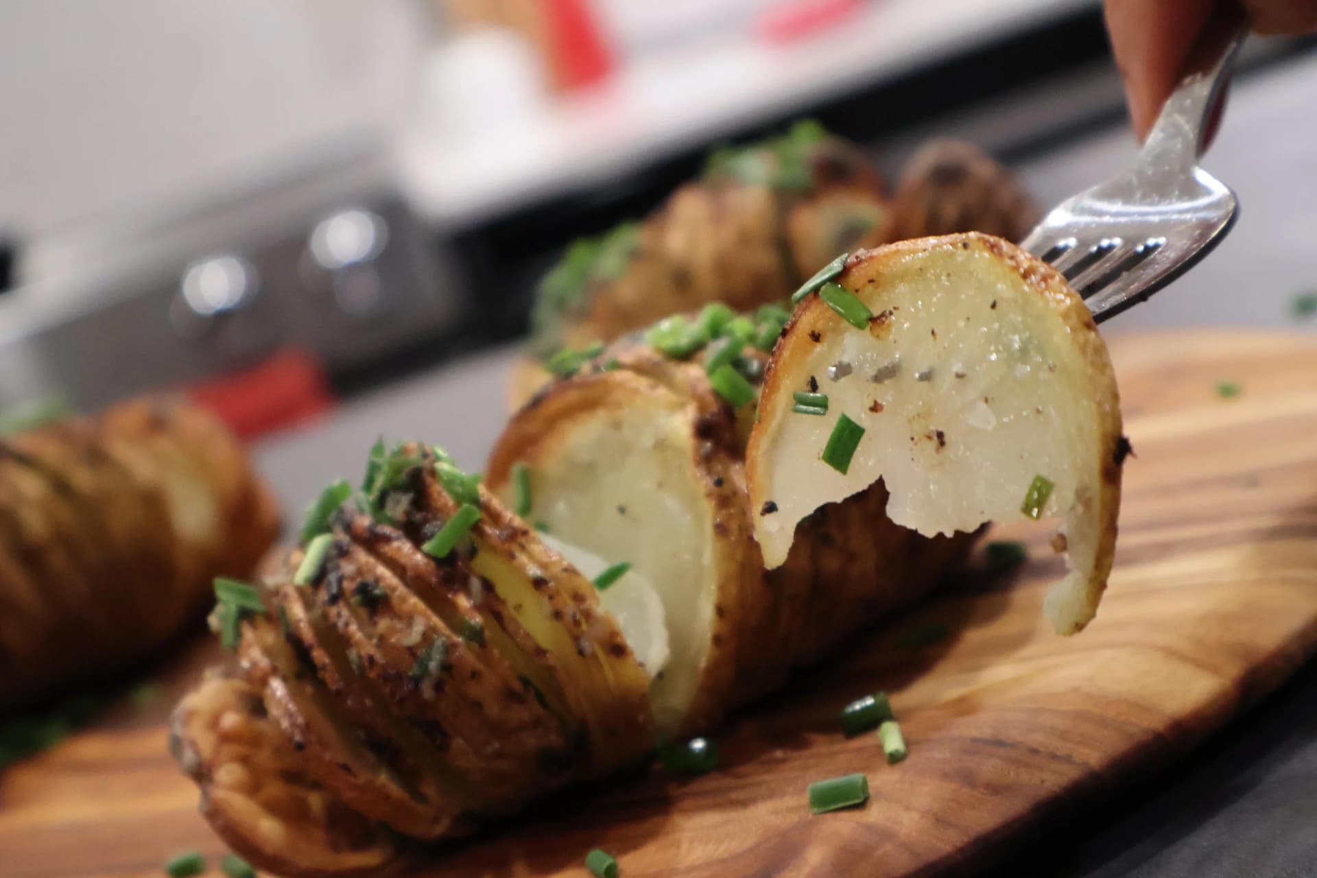 Hasselback potatoes on a wooden board garnished with chopped chives, being lifted with a fork to show the sliced layers.