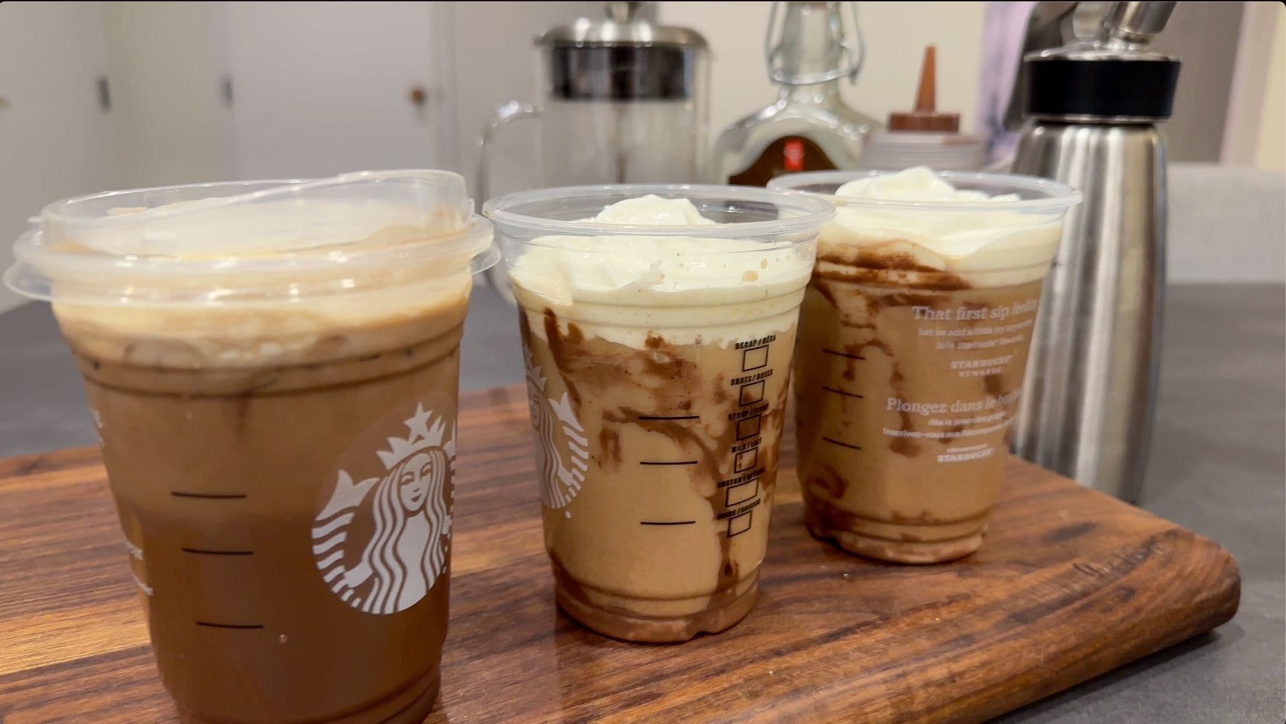 Three iced mochas with whipped cream on a wooden board, next to kitchen items.