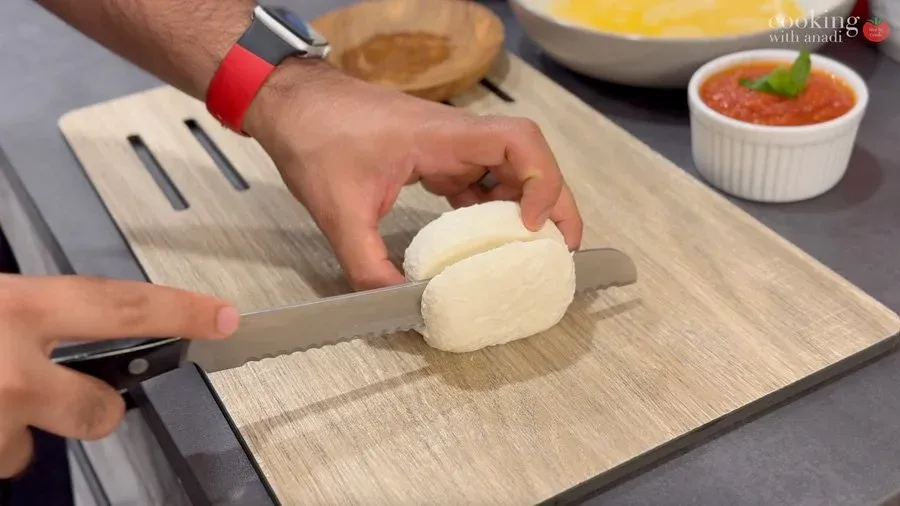 Slicing fresh mozzarella into thin rounds on a wooden cutting board for Mozzarella in Carrozza sandwich preparation.