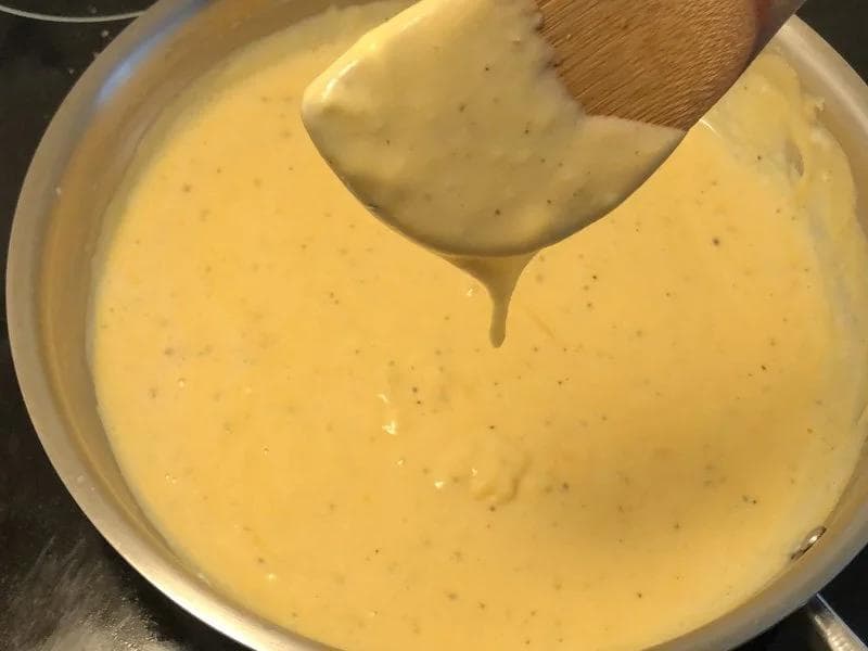 Creamy cheddar and mozzarella cheese sauce coating a wooden spoon over a pot of béchamel.