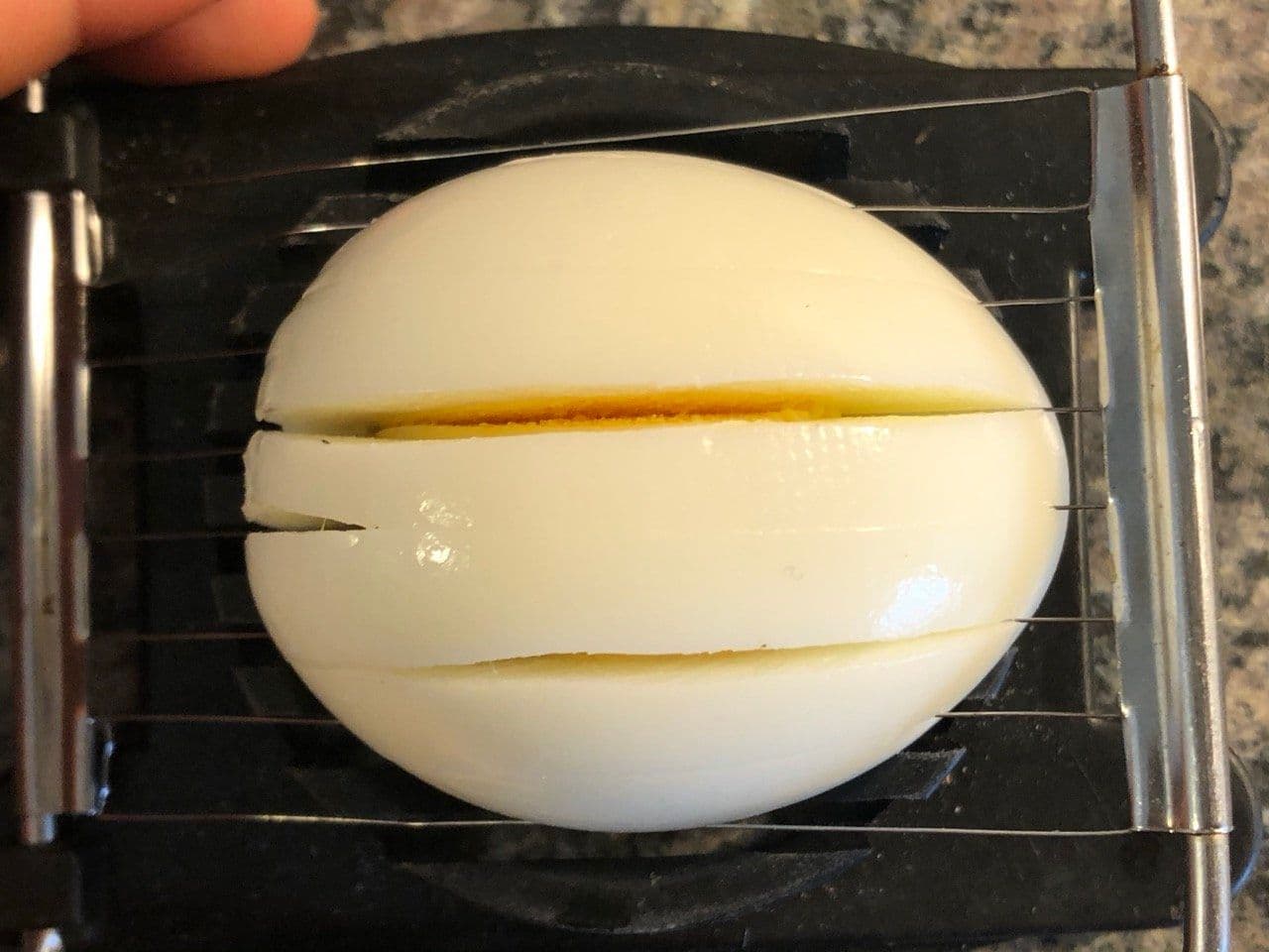 Hard-boiled egg being sliced into rounds using a metal egg slicer, revealing the yellow yolk inside.