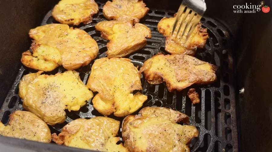 Golden-brown smashed potatoes cooking in an air fryer basket, seasoned with black pepper and partially melted cheese.