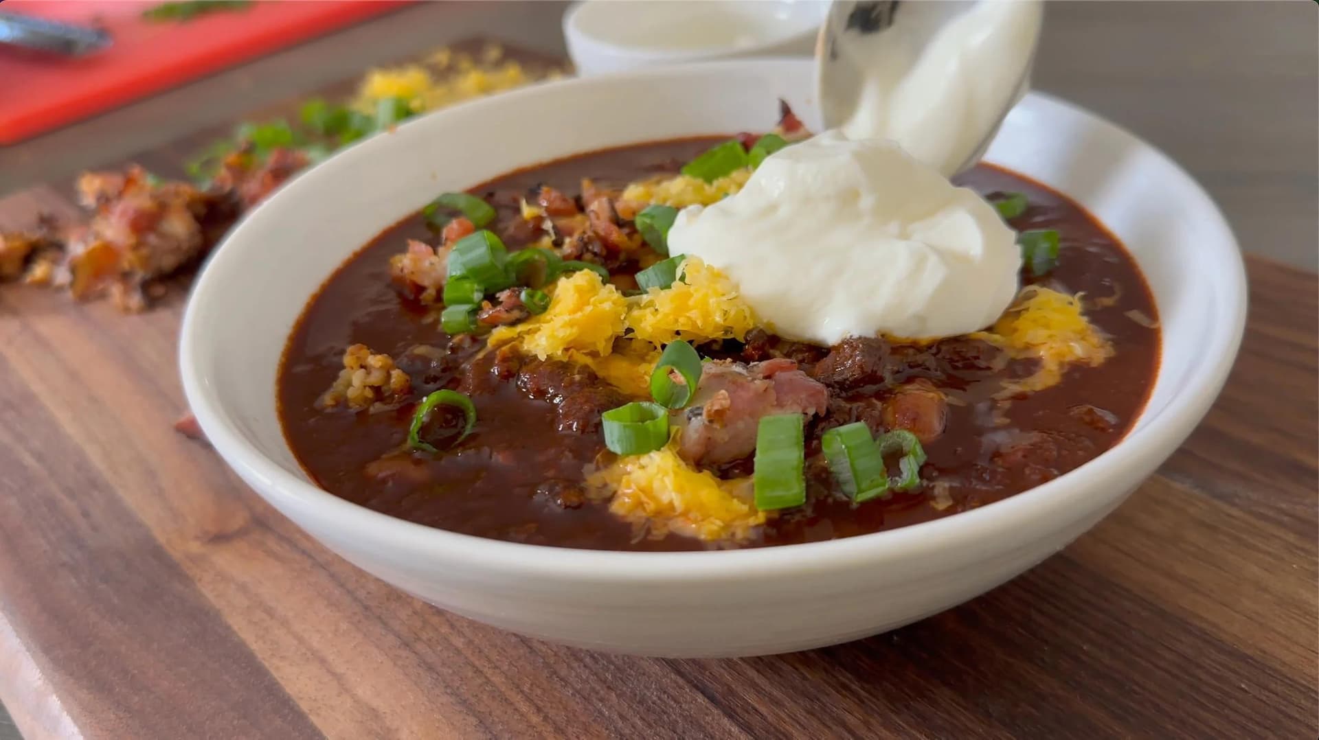 Texas chili topped with sour cream, shredded cheddar, green onions, and crispy bacon in a white bowl.