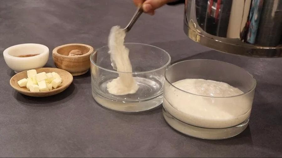 Pouring creamy rice pudding mixture into a glass bowl, with butter cubes and toppings arranged nearby on a dark counter.