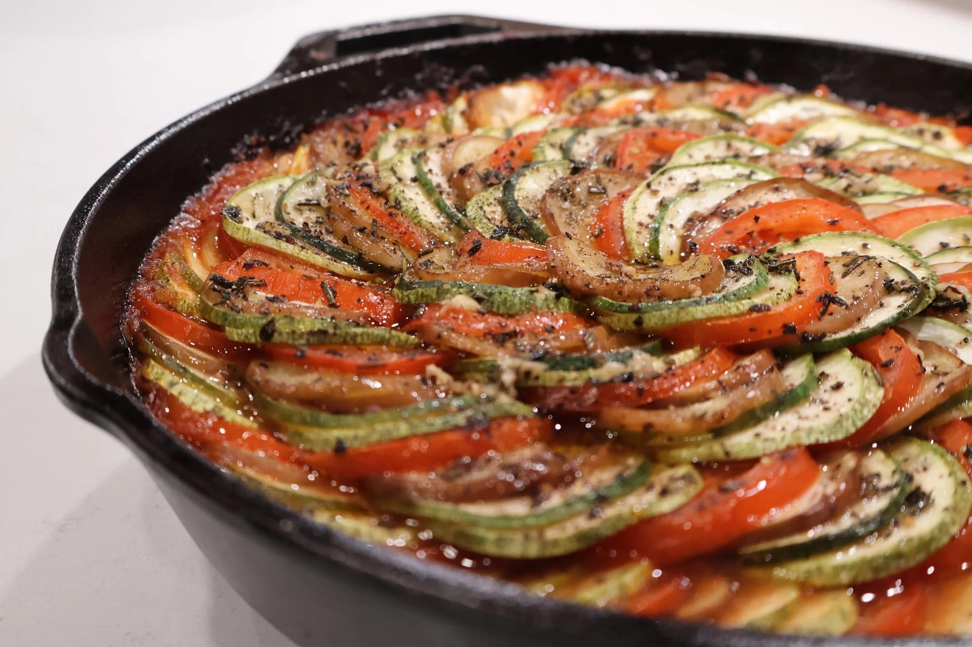 Ratatouille (confit byaldi) with overlapping zucchini, tomato, and onion slices in a cast iron skillet, seasoned with herbs.