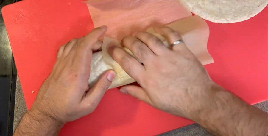 Hands wrapping a filled tortilla in parchment paper on a red surface.