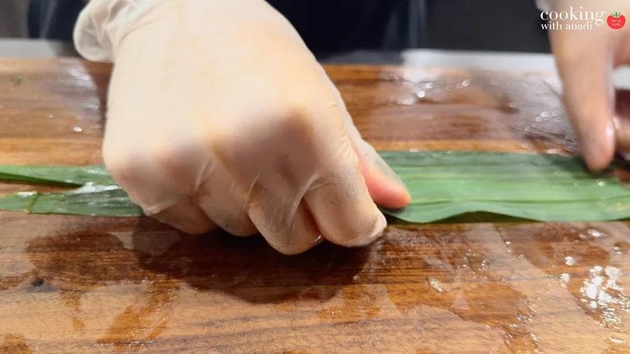 Gloved hand pressing and straightening fresh pandan leaves flat on a wooden cutting board.