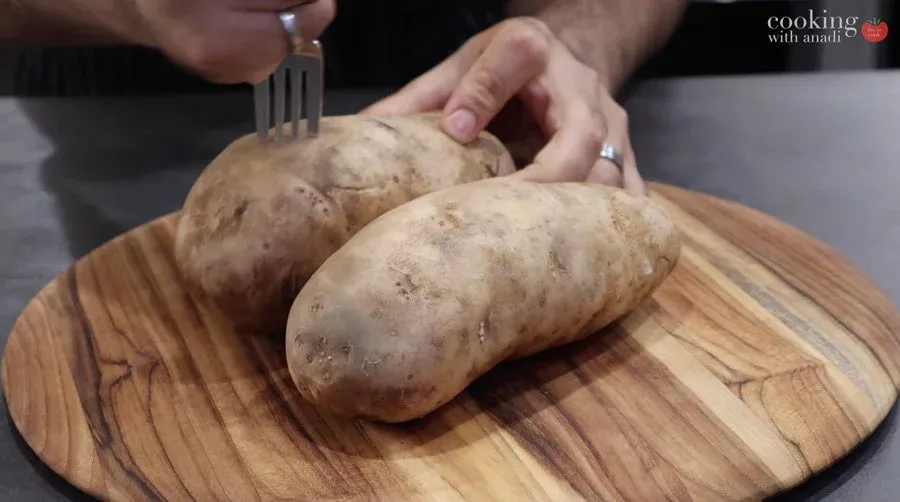 Two russet potatoes on a wooden board being pricked with a fork before baking.