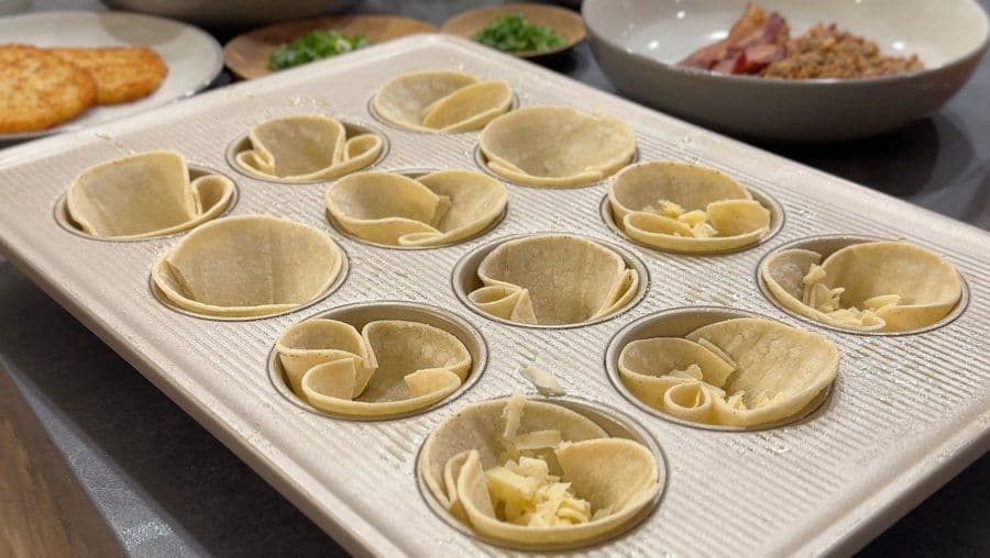Twelve tortilla cups pressed into a muffin tin with various vegetarian fillings like cheese and vegetables visible inside.