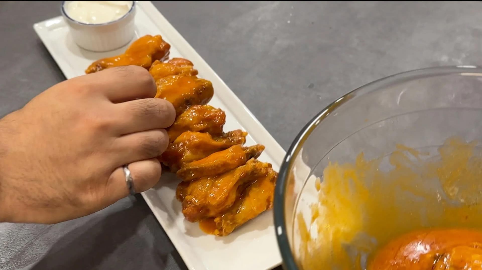 Hand holding a crispy fried buffalo chicken wing from a white plate, with hot sauce in a glass bowl nearby.