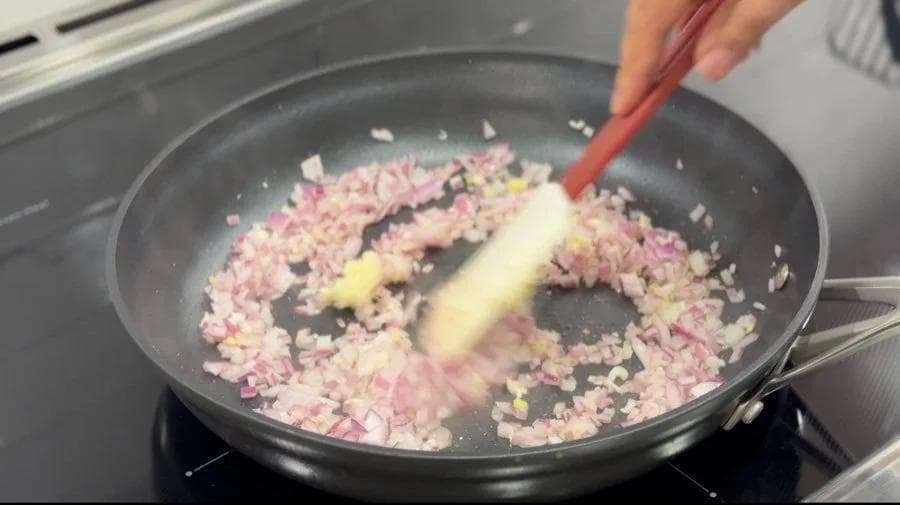 Diced red onions and garlic being sautéed in a dark pan with a spatula, early stage of refried beans preparation.