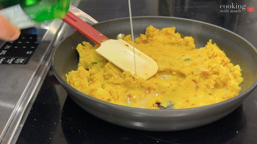Butter being added to a pan of cooked golden yellow potatoes with onions and spices using a red spatula.