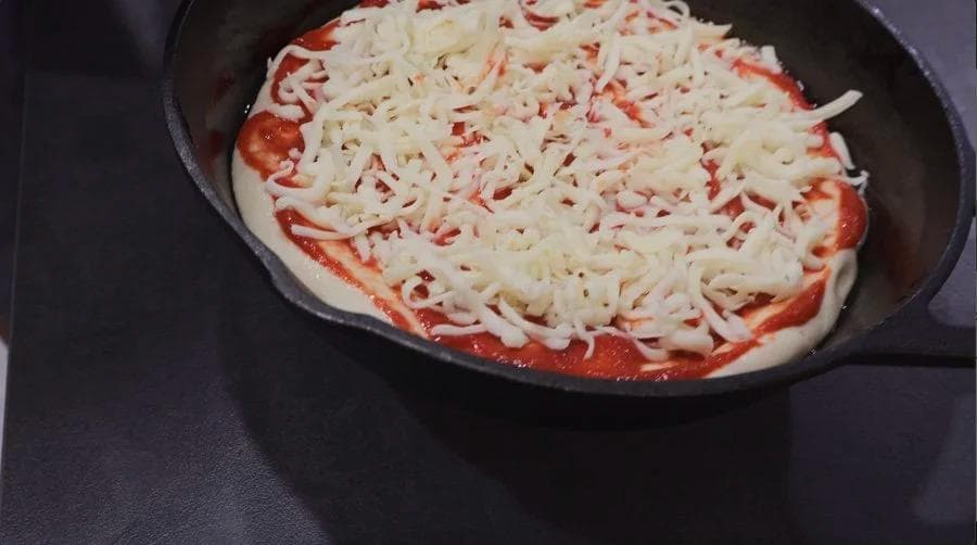 Cast iron skillet with pizza dough, red sauce, and melted mozzarella cheese ready for baking.