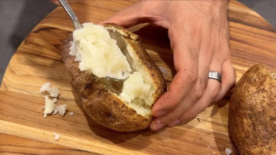 Hand scooping fluffy potato interior from halved baked potato shell on wooden board.