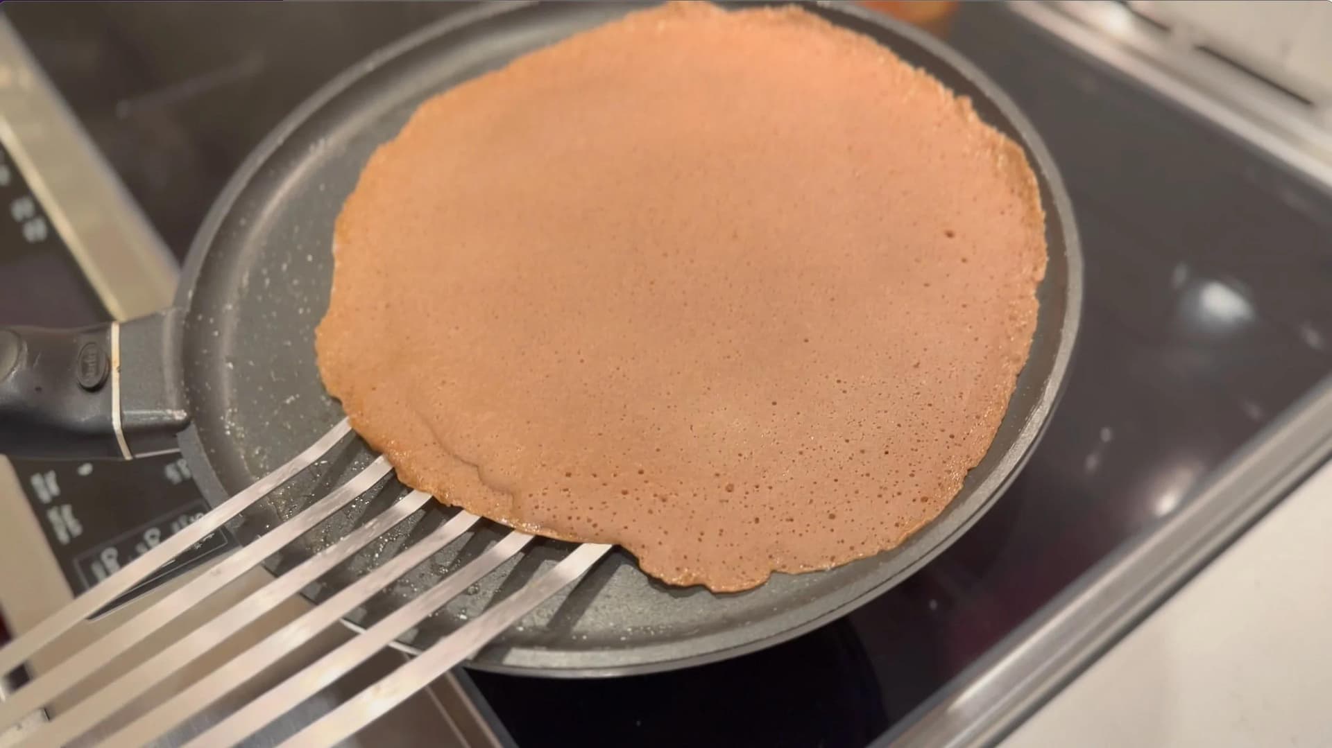 Golden-brown gluten-free chocolate crepe cooking in a non-stick pan, being flipped with a spatula.