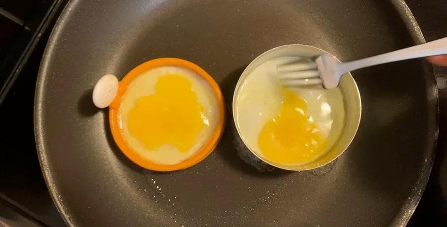Two eggs cooking in silicone rings on a skillet, one cracked and one whisked, for homemade breakfast sandwiches.