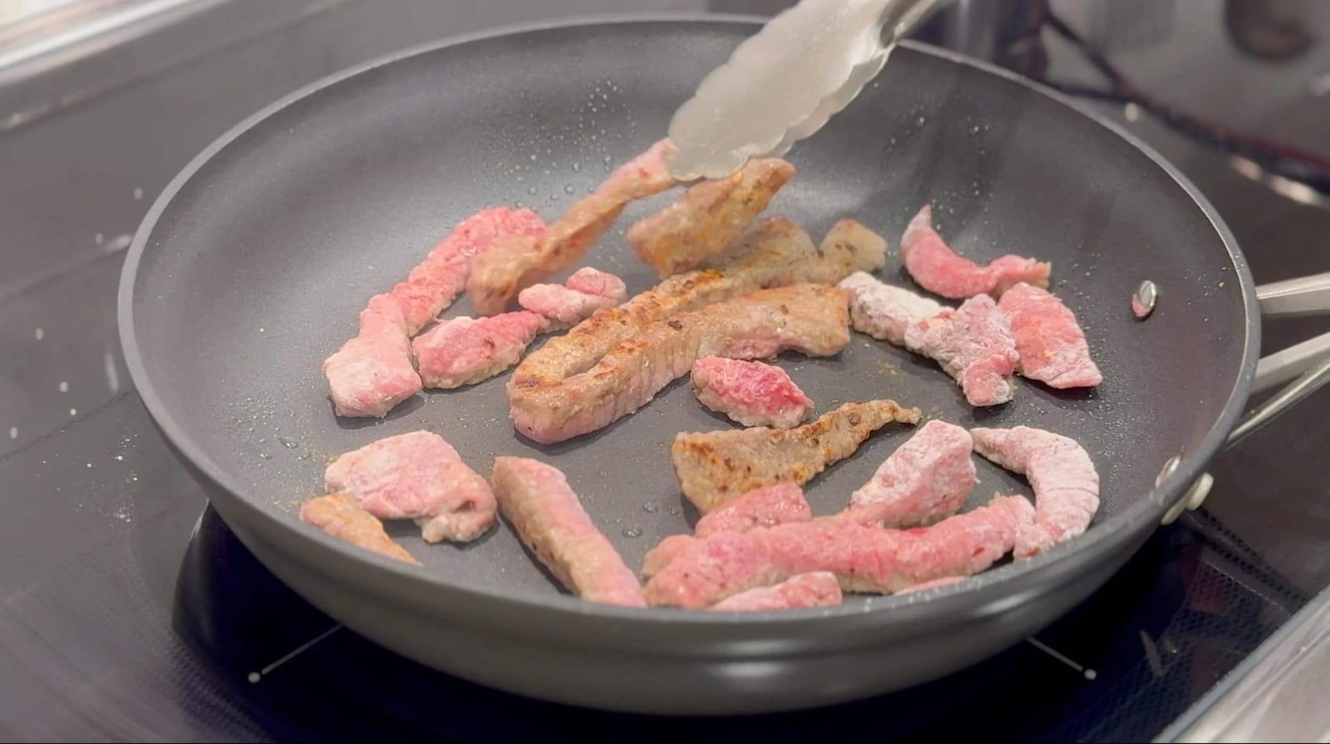 Breaded beef strips cooking in a pan, partially browned with pink centers, being flipped with a spatula.