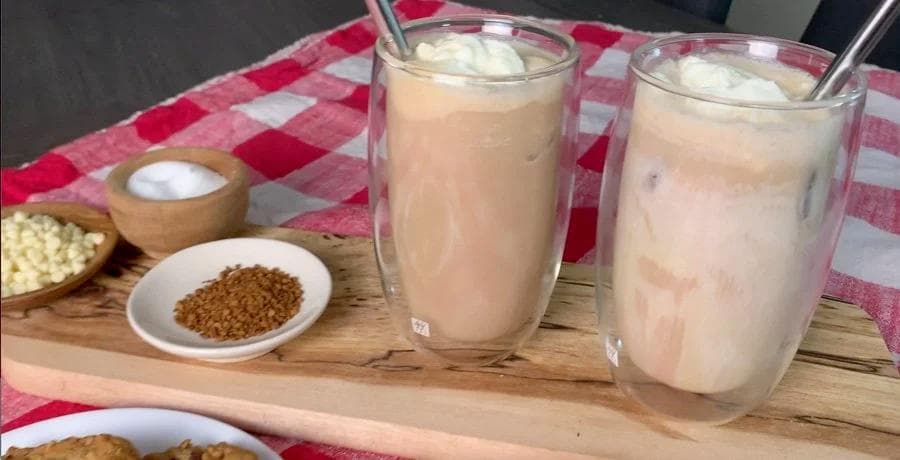 Two glasses of iced white chocolate mocha with whipped cream and ice, alongside ingredients in small bowls on a wooden board.