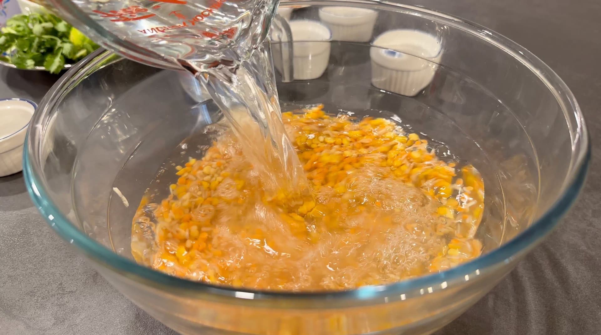 Liquid being poured into a glass bowl containing soaked lentils and rice with turmeric for haleem preparation.